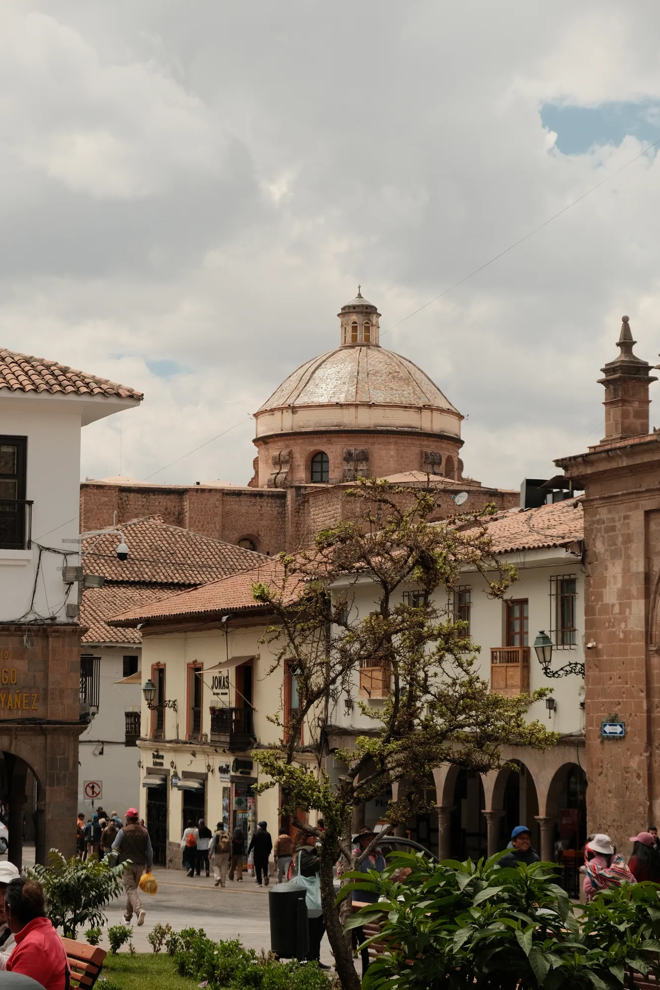 A colonial church dome rises over Plaza de Armas, seen past terracotta rooftops and a garden in central Cusco.