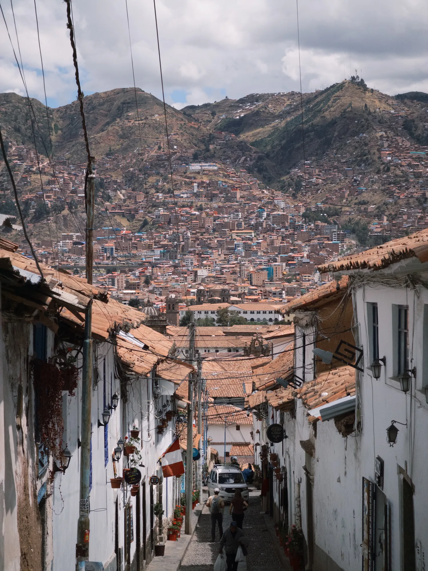 A steep cobblestone street in Cusco's San Blas neighborhood, framed by white-walled houses, looking down over the city and mountains beyond.