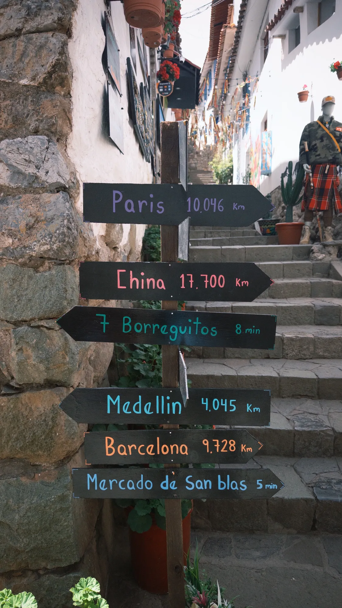 A hand-painted signpost on a San Blas staircase pointing to Paris, China, Barcelona, and the Mercado de San Blas, with distances marked in kilometers.