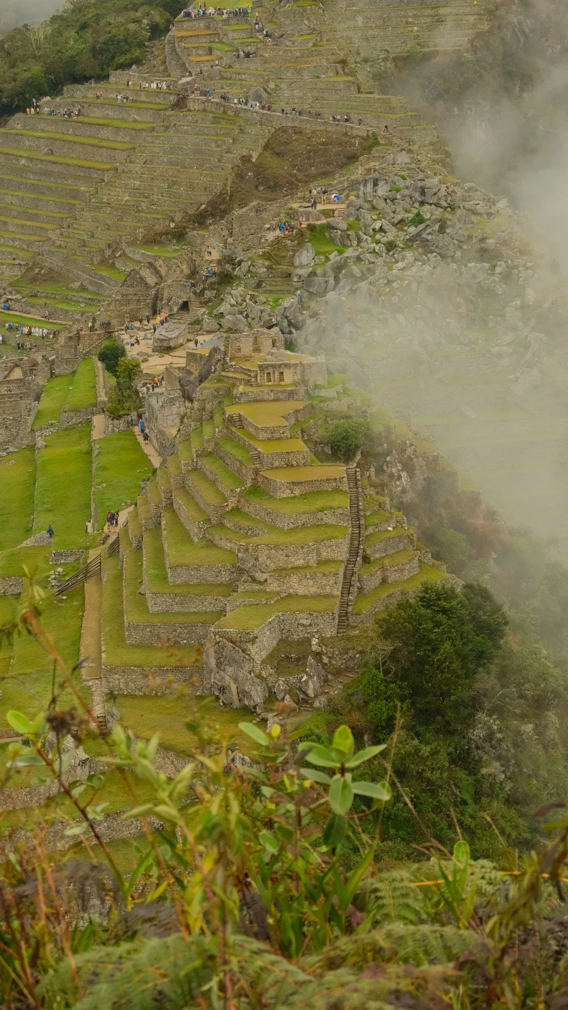 The classic view of Machu Picchu: stone terraces and ruins along the mountain ridge, cloud drifting past the cliff edge.