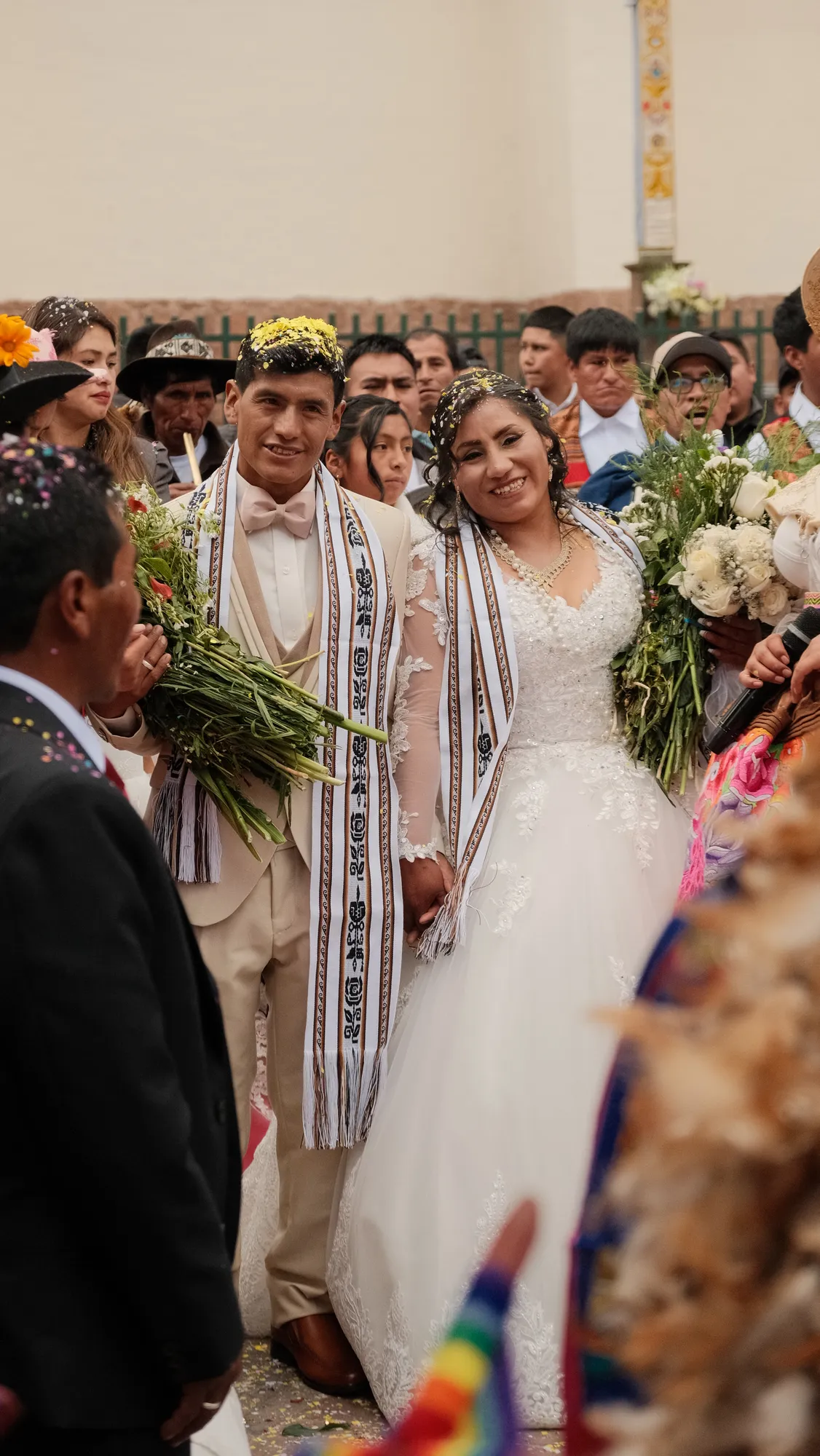A newly married couple in traditional Andean woven sashes and floral confetti hold hands outside a church in Pisac, surrounded by wedding guests.