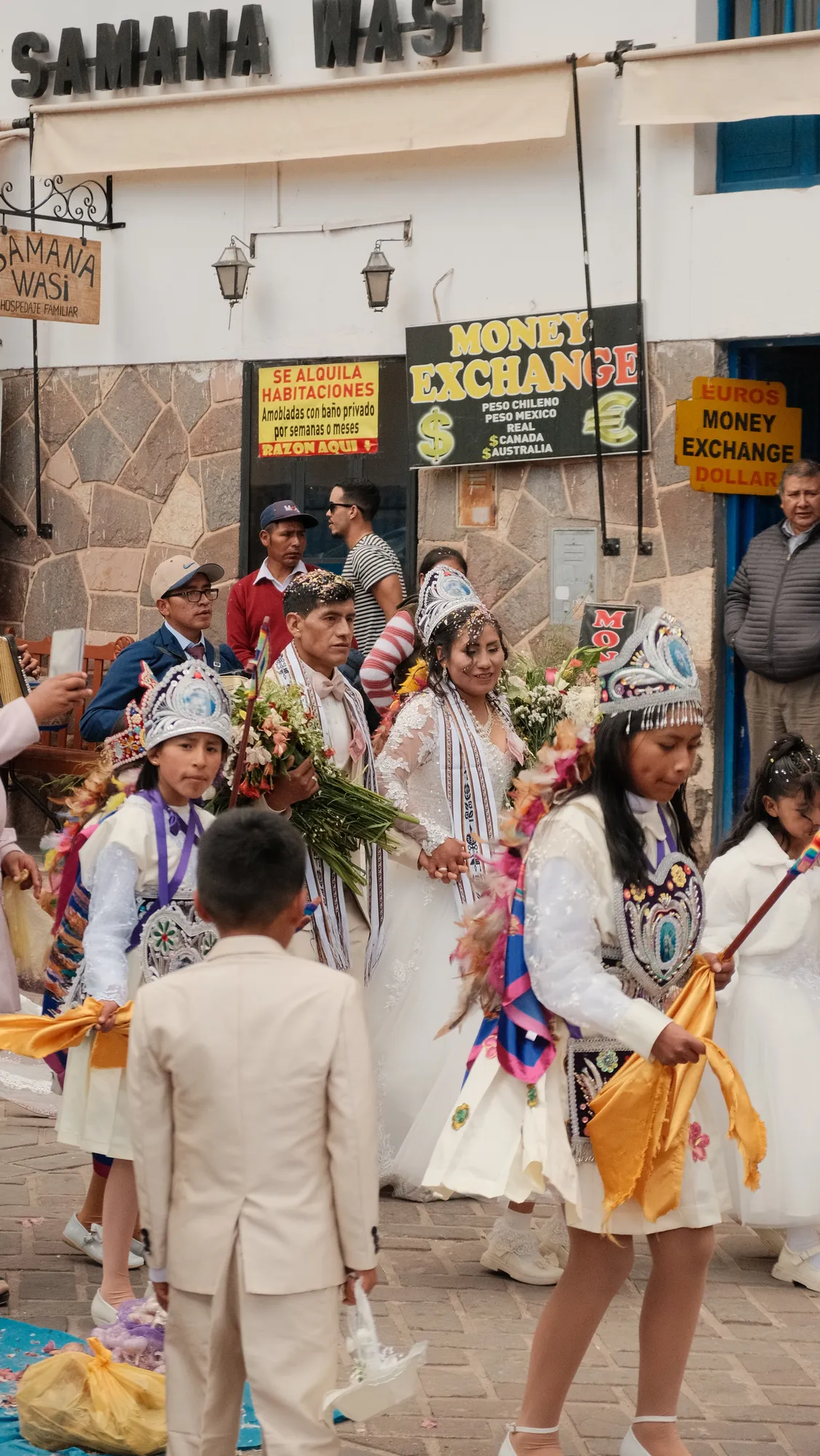 The wedding party continues through Pisac's plaza past market stalls and umbrellas.