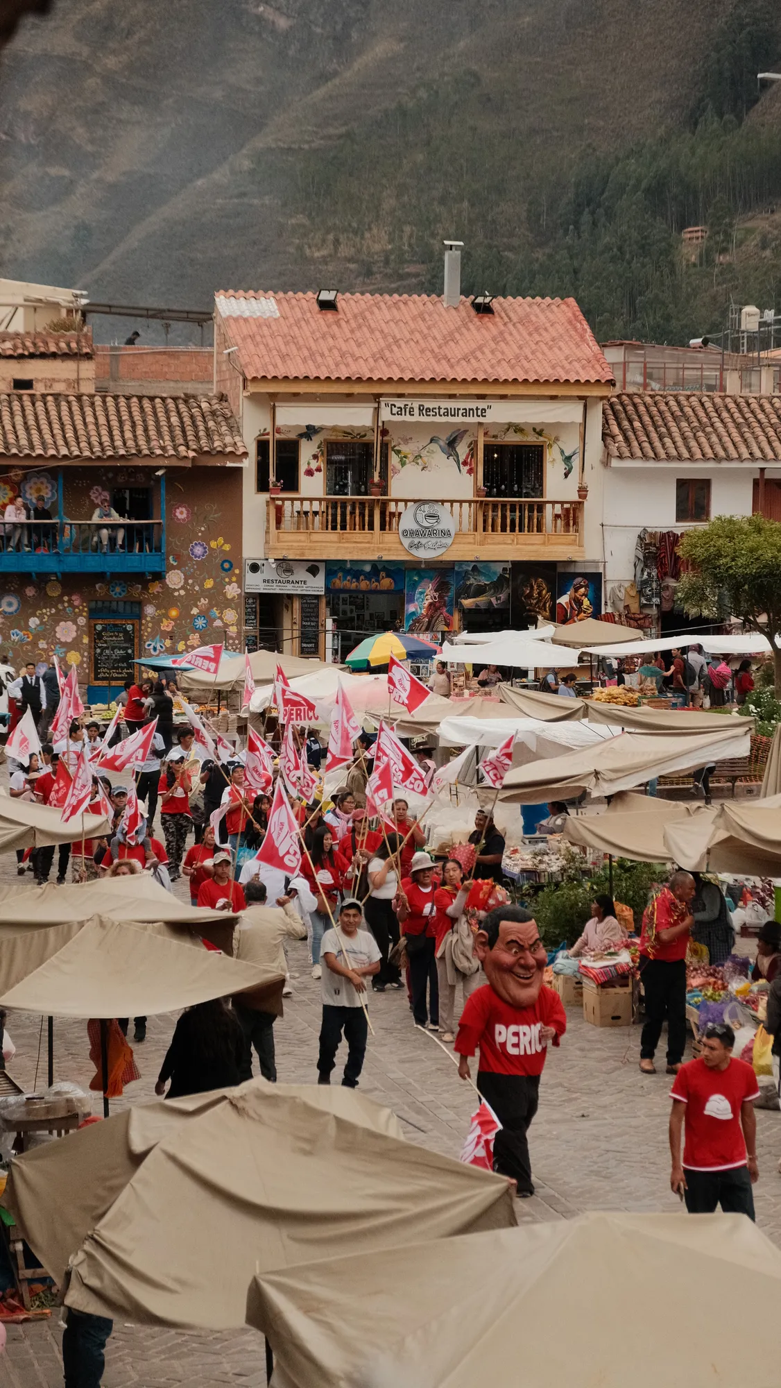 A parade with red flags and a costumed mascot moves past market stalls in Pisac's plaza, mountains behind the town.