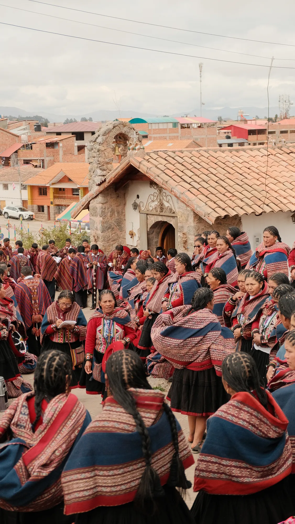 A large community gathering fills the square outside a small stone chapel in Chinchero, women in red-and-navy woven shawls with braided hair, the town and mountains behind.
